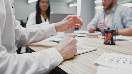 Hand of unknown employee who writing down information into a notepad, diverse colleagues having business meeting in office