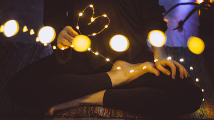 Woman seated cross-legged with bokeh Christmas lights in a heart shape. Concept: love and magic in...