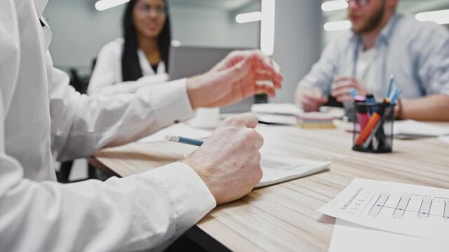 Hand of unknown employee who writing down information into a notepad, diverse colleagues having business meeting in office