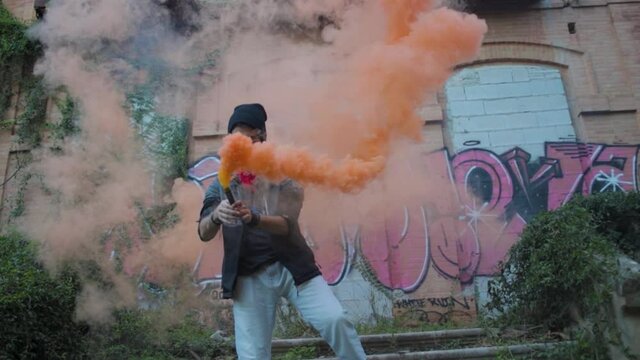 hombre con bomba de humo naranja en lugar abandonado con la cara tapada por un pa&ntilde;uelo