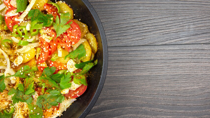 cooked fried potatoes with herbs, spices and vegetables in a black pan on a wooden table. copy space