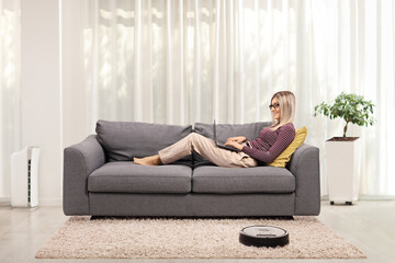 Young woman working on a laptop and laying on a sofa at home with a robotic vacuum cleaner