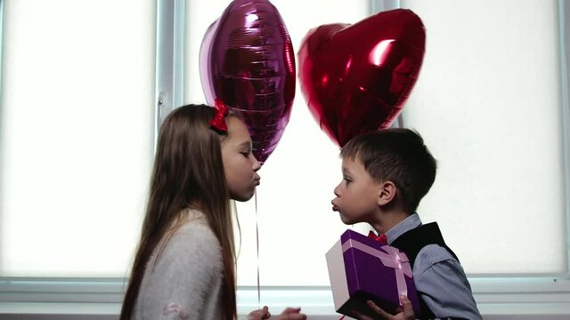 An attractive, elegant sister holds a gift box and a balloon behind her back, and then gives it to her brother. Kids smack each other on the lips, Valentine's Day