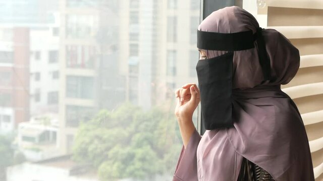 young muslim women covering face looking through window indoor