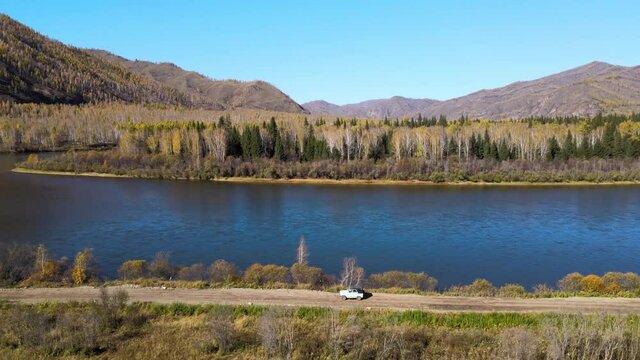 The car drives along a rural road along a blue river with mountains in the background. UAZ goes from right to left. Nice panoramic view from the quadcopter. Russia. Tyva Republic. The Yenisei River. S