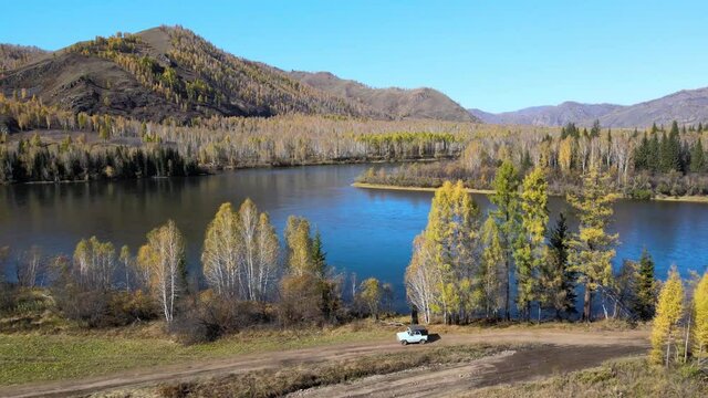 The car drives along a rural road along a blue river with mountains in the background. UAZ goes from right to left. Nice panoramic view from the quadcopter. Russia. Tyva Republic. The Yenisei River. S
