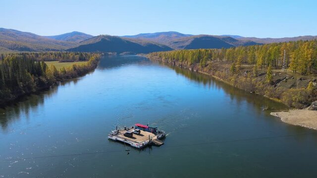 A pontoon ferry crossing the blue Yenisei River in the Tyva Republic, Russia. Two cars are ferrying to the other side of the river. In the distance you can see the mountains and on the shore there is 