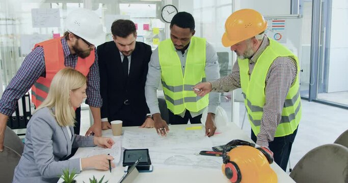 Attractive confident professional multiracial team of engineers of building company analyzing construction drawing with their successful high-skilled bearded ceo during meeting in boardroom