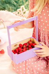 girl on a picnic in a spring park. female hands hold a purple wooden basket with strawberries.