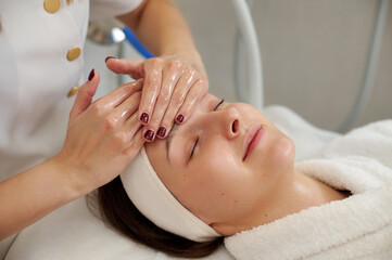 Fototapeta na wymiar Hands of cosmetology specialist making facial massage for pretty young woman in SPA salon