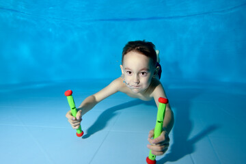 Cute boy fun dives to the bottom of the pool. He smiles and looks into the camera frame. Fun dives under the water. Active happy child. Swimming classes. Bodily exercises. Healthy lifestyle