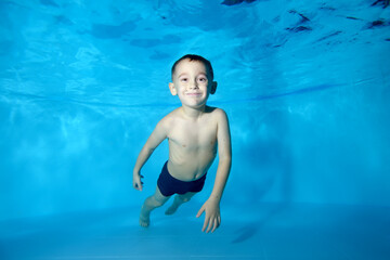 Photo of a sporty boy at the bottom of a children's pool. He smiles and looks into the camera frame. Fun dives underwater. Active happy child. Swimming classes. Bodily exercises. Healthy lifestyle