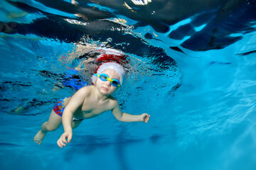 Portrait of a little boy in a Christmas hat and glasses in a pool under water. He looks at the camera. Baby learns to dive. Swimming lessons with a child. Healthy lifestyle. Horizontal orientation © alexbard