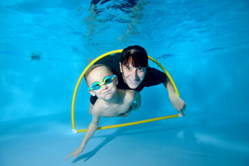 A happy toddler and a female trainer swim together underwater through a Hoop. They smile and look...