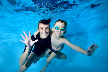 Portrait of a beautiful boy and his coach underwater. They smile and look at the camera. Active happy child. Swimming lessons for small children. Healthy lifestyle
