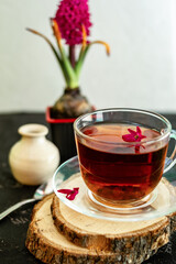 A cup of tea in a transparent cup with spring hyacinth flower on a black background. The mug stands on a wooden frame, next to it lies a metal spoon and a white vase. Copy space