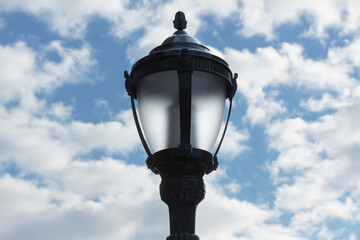 Fototapeta na wymiar an unlit street lamp made of black cast iron with sculptural details against a cloudy, blue sky during the day