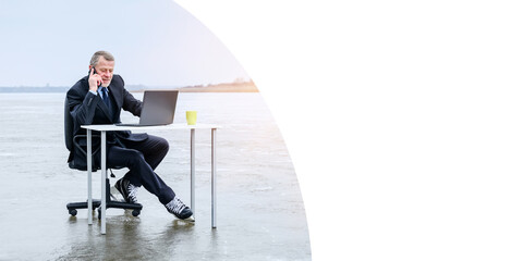 gray-bearded smiling senior businessman in suit and skates, smiling while talking on smartphone, works with laptop on table in the middle of a frozen lake. Banner, copy space, remote work concept