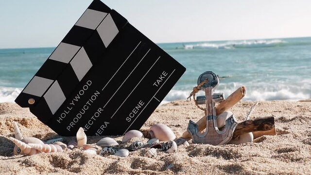 Photo of a movie clapperboard and a set of wooden anchor and sea shells with an out of focus background of surfers.