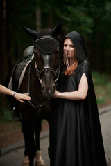 Naklejka premium Young woman in hat and black cloak with a horse in the forest