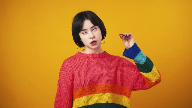 Young bored woman in red sweater with rainbow sleeves showing blah gesture and rolling up her eyes, orange background