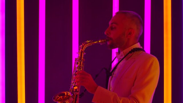 A musician in a white suit is masterly playing the golden saxophone. Side view of a man in the studio against the background of multicolored neon pipes. Slow motion, close up.