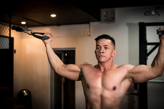 A fit and toned asian man with a serious and determined look while working out, doing standing cable double bicep curls at the gym.