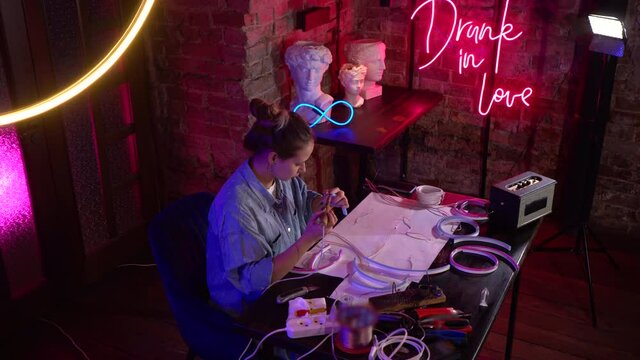 A young beautiful girl makes neon signs in a workshop. A talented artist professionally works with a soldering iron in an ultraviolet studio. Thick smoke rises.