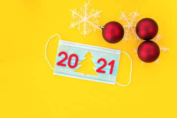 Medical mask with cut-out silhouette of Christmas tree and red numbers 2021 with snowflakes and red Christmas balls on yellow background.New Year is coming.2021 is coming.New Year concept.