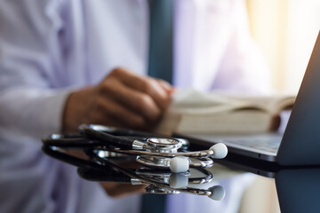 Doctor man in white lab coat reading text book and work on laptop computer with medical stethoscope on the office desk at clinic. Online medical education concept.