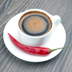 Black coffee in a white cup with red hot pepper on a plate. Spicy food and strong drink.