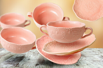 Set of pink empty porcelain teacups with saucers flying over wooden table surface
