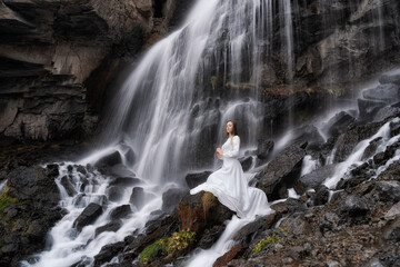 A girl in a white dress sits by a waterfall