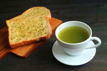 Closeup a Cup of Hot Green Tea with Blurry Garlic Butter Toasts on Black Wooden Table