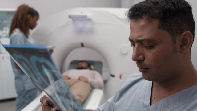 PAN close up of Arab male doctor reading x-ray of lungs while black female tech talking to patient lying in CT scan machine