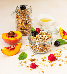 Homemade crunchy granola in glass jar, peaches, berries, honey and mint, light concrete background....