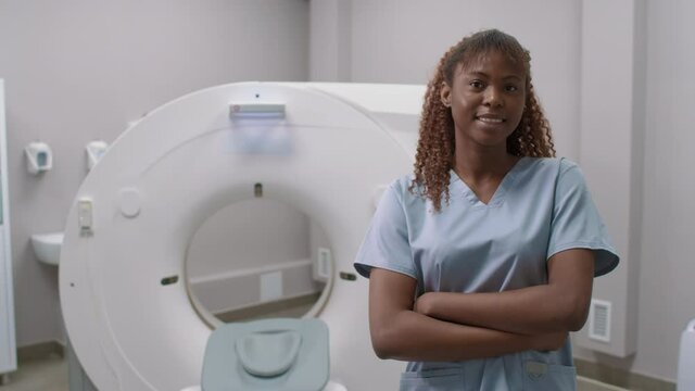 Panning portrait shot of happy female radiologist in scrubs standing with her arms crossed in CT scan room in hospital and smiling for camera