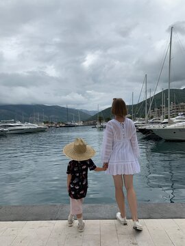 A girl in a white dress together with a little girl standing with their backs by the sea. Mother with a daughter. Walk in the marina. Cloudy weather. iPhone photo.