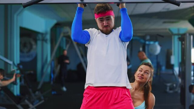 Caucasian fitness woman training weak bearded man training in the gym exercising pull ups going hard intense workout. Funny couple. Workout.