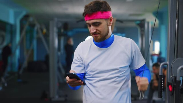 Funny bearded caucasian guy using smartphone while exercising lazy beginner training at the gym. Nomobphobia. Mobile phone addiction.