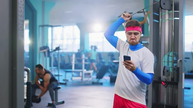 Funny caucasian weak athlete exercising on gym equipment getting motivated by fitness pictures on smartphone using device indoors. Fitness parody. Humor concept.