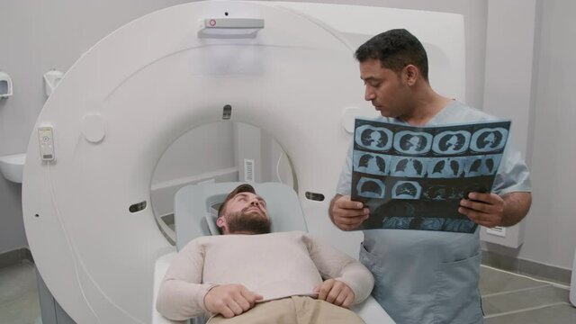 Handheld tracking of Arab male radiologist holding chest x-ray and talking to patient lying on treatment couch in CT scan
