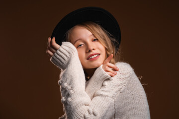 Charming blonde teen girl in black hat posing in studio on brown background