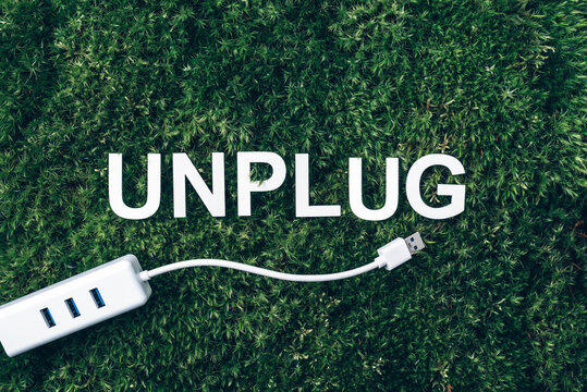 Word Unplug, white internet usb adapter on moss, green grass background. Top view. Copy space. Banner. Biophilia concept. Nature backdrop