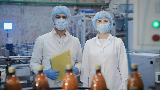 Happy colleagues in medical masks and uniform give five standing near conveyor with juice bottles in contemporary plant workshop