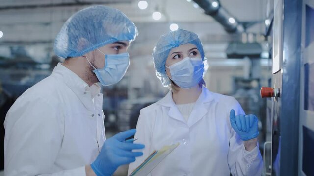 Skilled inspectors in white coats and disposable masks check data of production line equipment in contemporary plant workshop