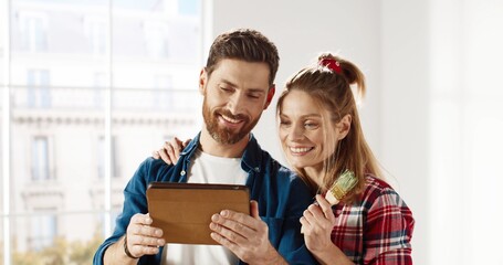 Close up portrait of cheerful young married couple man and woman stand in new apartment, tapping and browsing online on tablet, choosing and discussing new design of house. Home repair