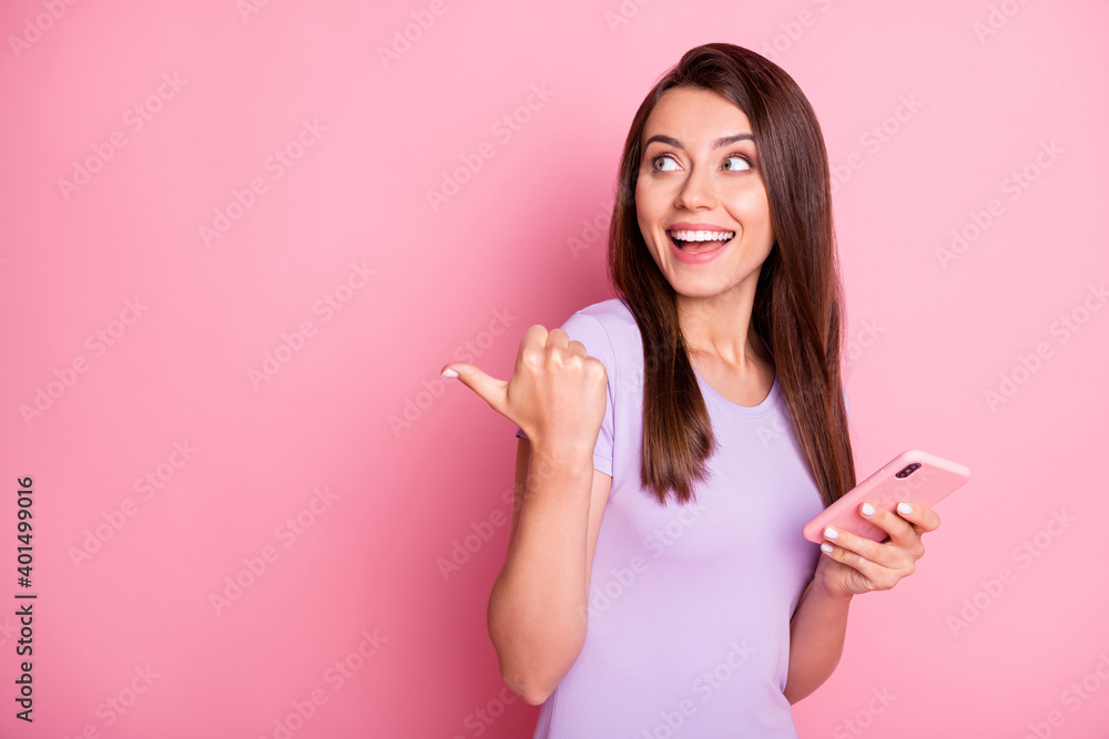 Poster Photo of funky excited young woman brown hair hold phone show pointing gesture look empty space dressed purple t-shirt isolated on pink color background