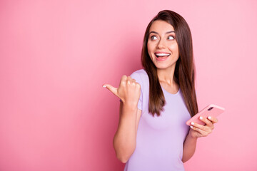 Photo of funky excited young woman brown hair hold phone show pointing gesture look empty space dressed purple t-shirt isolated on pink color background