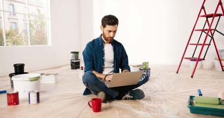 Caucasian happy bearded young handsome man sitting on floor in empty apartment during repair works...